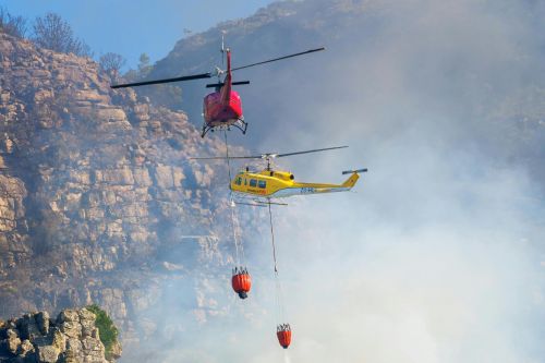 Two helicopters extinguishing a wildfire in a mountainous area, showcasing aerial firefighting efforts.