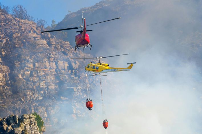 Two helicopters extinguishing a wildfire in a mountainous area, showcasing aerial firefighting efforts.
