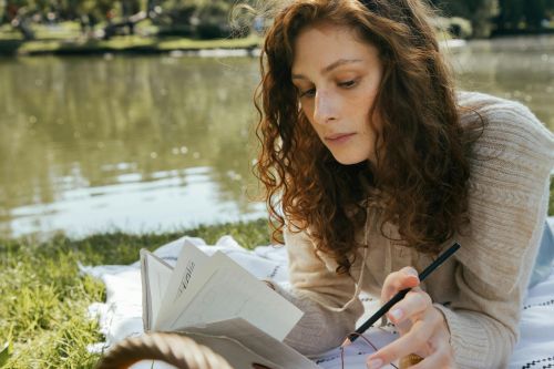 Woman with curly hair writing in a notebook outside by a serene lake on a sunny summer day.