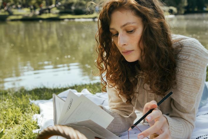 Woman with curly hair writing in a notebook outside by a serene lake on a sunny summer day.