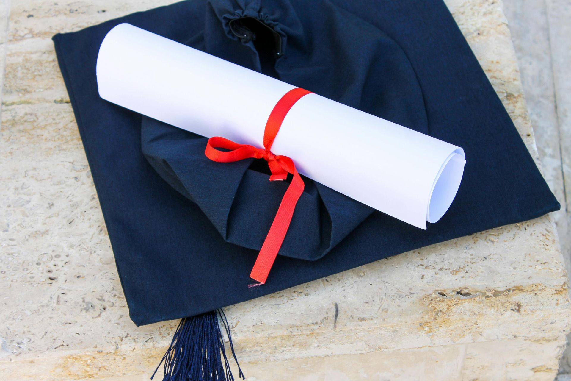 Graduation cap with diploma roll tied with red ribbon on concrete surface.